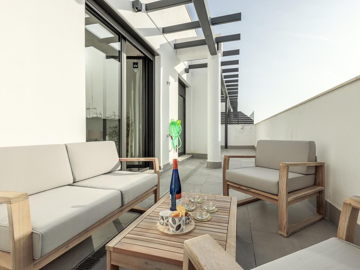 Et moderne udendørs opholdsområde med sofa og stole, et træbord med drikkevarer og glas, samt en glasdør der fører til en balkon.