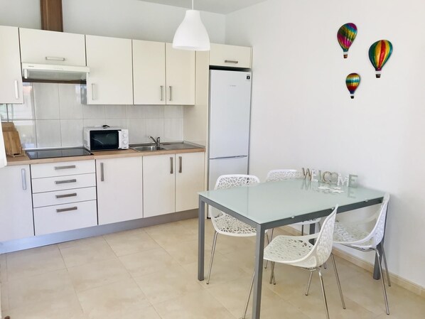 A modern kitchen with white cabinets, a microwave, and a refrigerator. There is a dining table with white chairs and a 'WELCOME' sign on the wall.