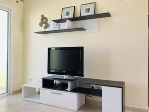 A modern living room with a flat-screen TV on a black stand, a white cabinet below, and two black shelves above holding framed pictures and decorative items.