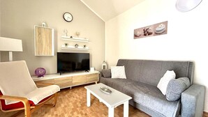 A living room with a grey sofa, a white coffee table, a TV on a wooden cabinet, a clock on the wall, and a small shelf with decorative items. - Rio Sol - Oasis of privacy (Costa Calma (incl. Urbanización Cañada d)