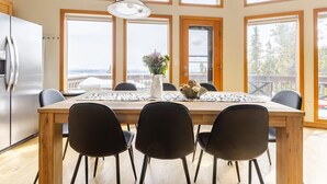 Une cuisine moderne avec une table en bois, des chaises noires, un réfrigérateur en acier inoxydable et de grandes fenêtres donnant sur l’extérieur.