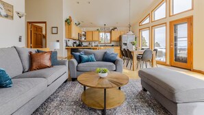 Un vaste espace de vie comprenant un canapé gris, une table basse ronde et une table à manger avec des chaises. On aperçoit, au fond, une cuisine avec des meubles en bois et une vue dégagée grâce à de grandes fenêtres.