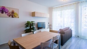 Una sala de estar moderna con una mesa de comedor de madera, sillas blancas, un sofá, un televisor y una planta.