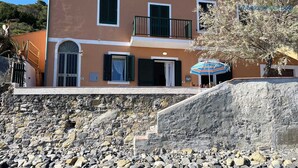 Un edificio costero con fachada de piedra, múltiples ventanas con contraventanas y un balcón con sombrilla a rayas.