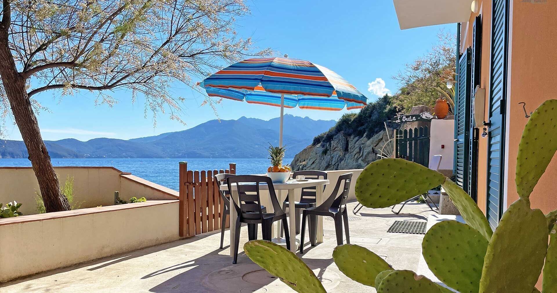 Un patio al aire libre con una mesa y sillas, un colorido parasol a rayas y vistas al mar y las montañas.