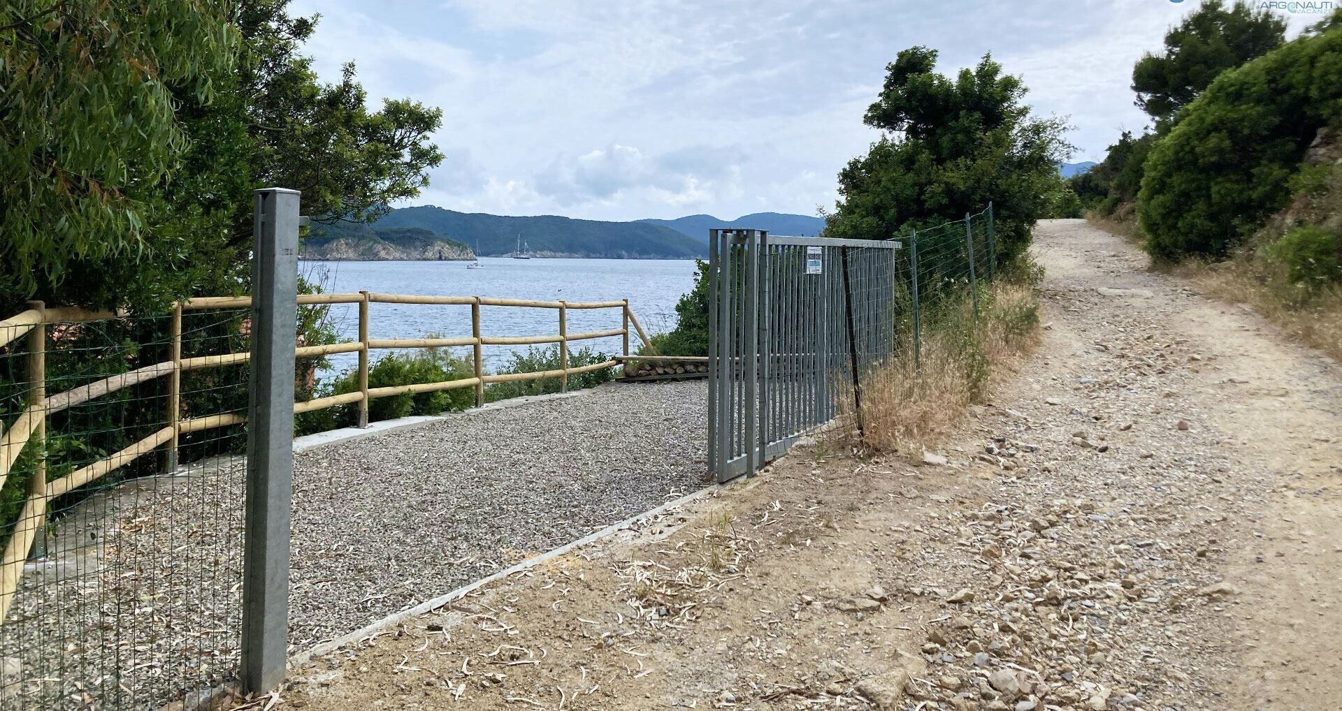 Ein Kiesweg führt zu einem Holzzaun und einem Metalltor, im Hintergrund ein Gewässer und Hügel.