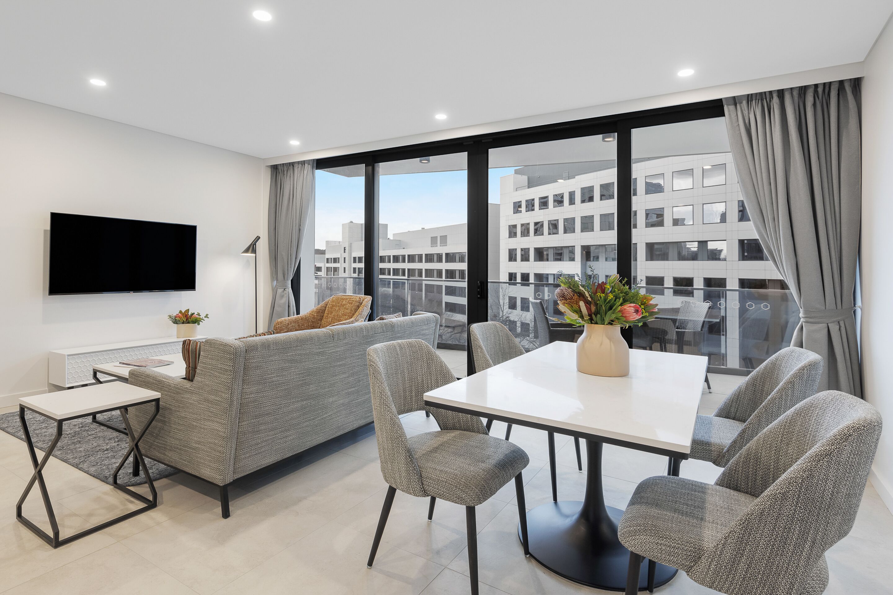 A modern living room with a grey sofa, a white dining table, and grey chairs. There is a flat-screen TV mounted on the wall and a potted plant on the table.