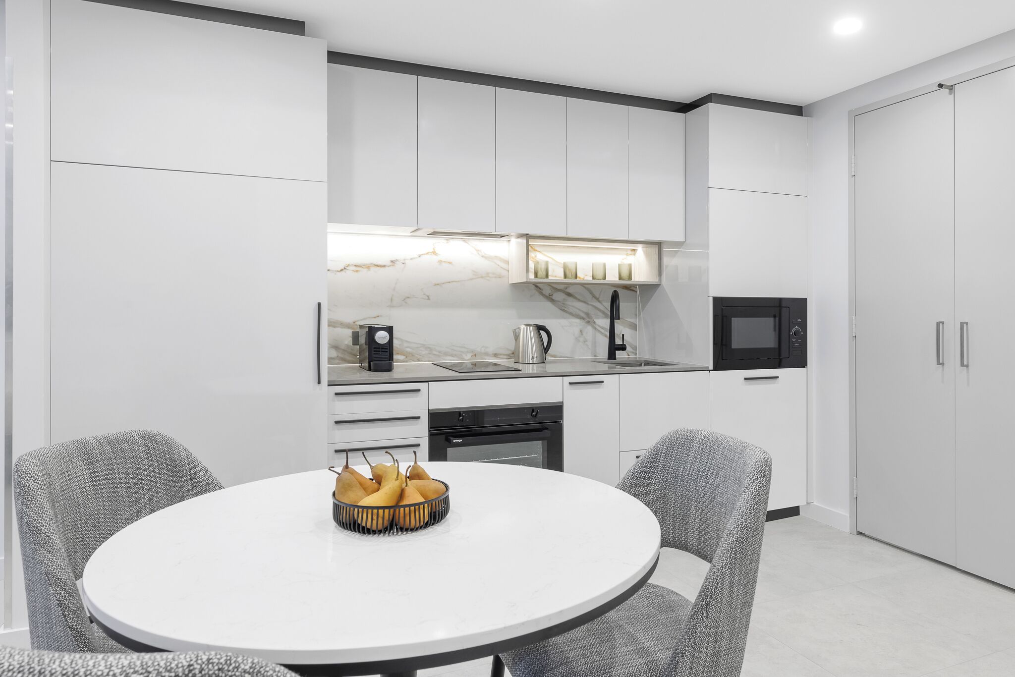 Una cocina moderna con armarios blancos, encimera de mármol y una zona de comedor con una mesa redonda y dos sillas.