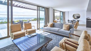Una sala de estar moderna con un sofá, sillones y una mesita de centro. Amplias ventanas ofrecen vistas de la ciudad.