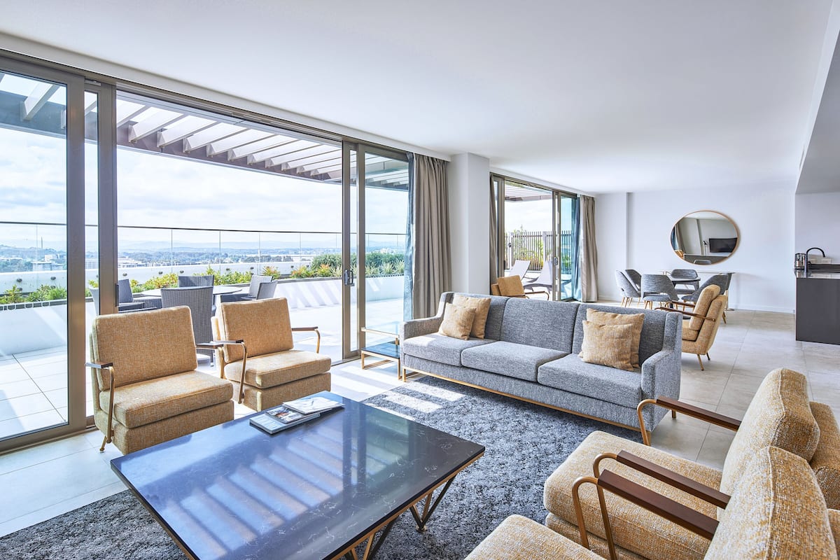 A modern living room with a sofa, armchairs, and a coffee table. Large windows offer a view of the city.
