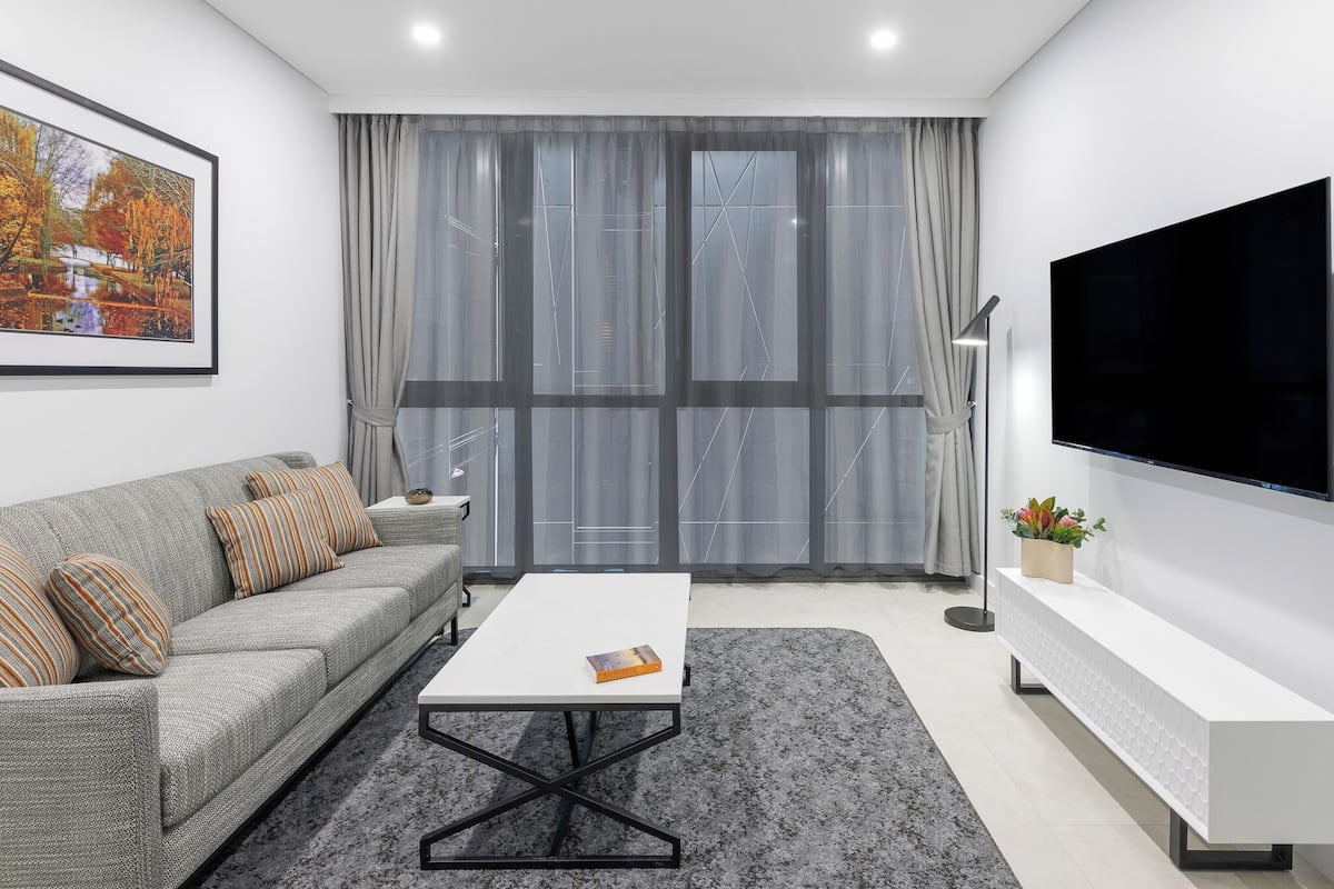A modern living room with a grey sofa, a white coffee table, a large flat-screen TV, and a grey patterned rug.