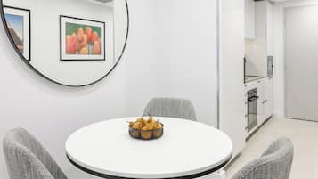 A modern dining area with a round table, grey upholstered chairs, and a fruit bowl. There are framed pictures on the wall and a kitchen area in the background.