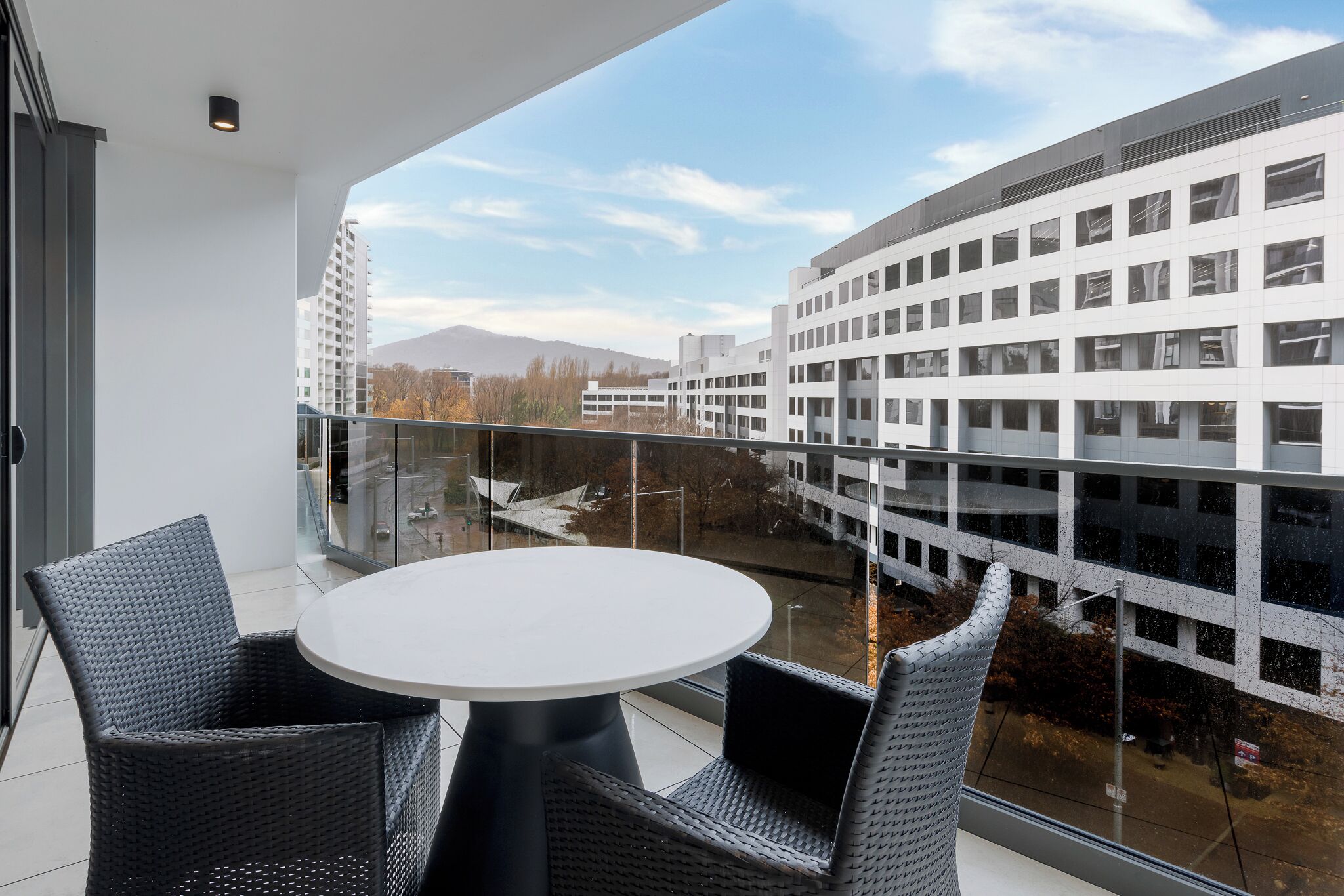 A balcony with a round table and two chairs, overlooking a modern building and a clear sky.