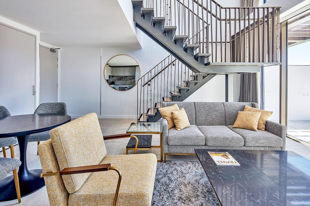 A modern living room with a grey sofa, a round mirror, and a staircase leading to an upper level.
