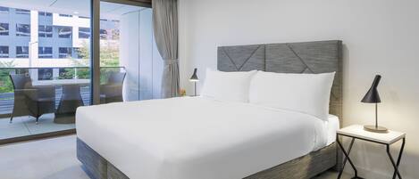 Habitación de hotel moderna con una cama grande, una mesita de noche pequeña con lámpara y vistas al balcón exterior.