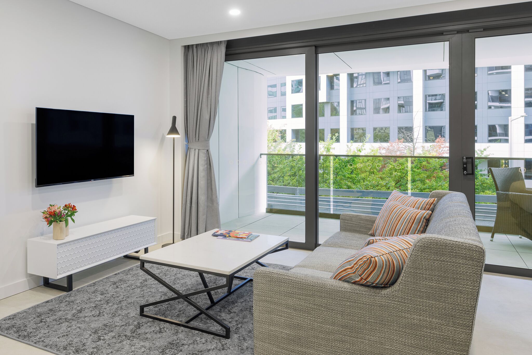 En moderne stue med et fladskærms-tv, et hvidt konsolbord med en vase med blomster, en grå sofa med stribede puder og en stor skydedør af glas, der fører ud til en balkon med udsigt over bygninger og grønne områder.