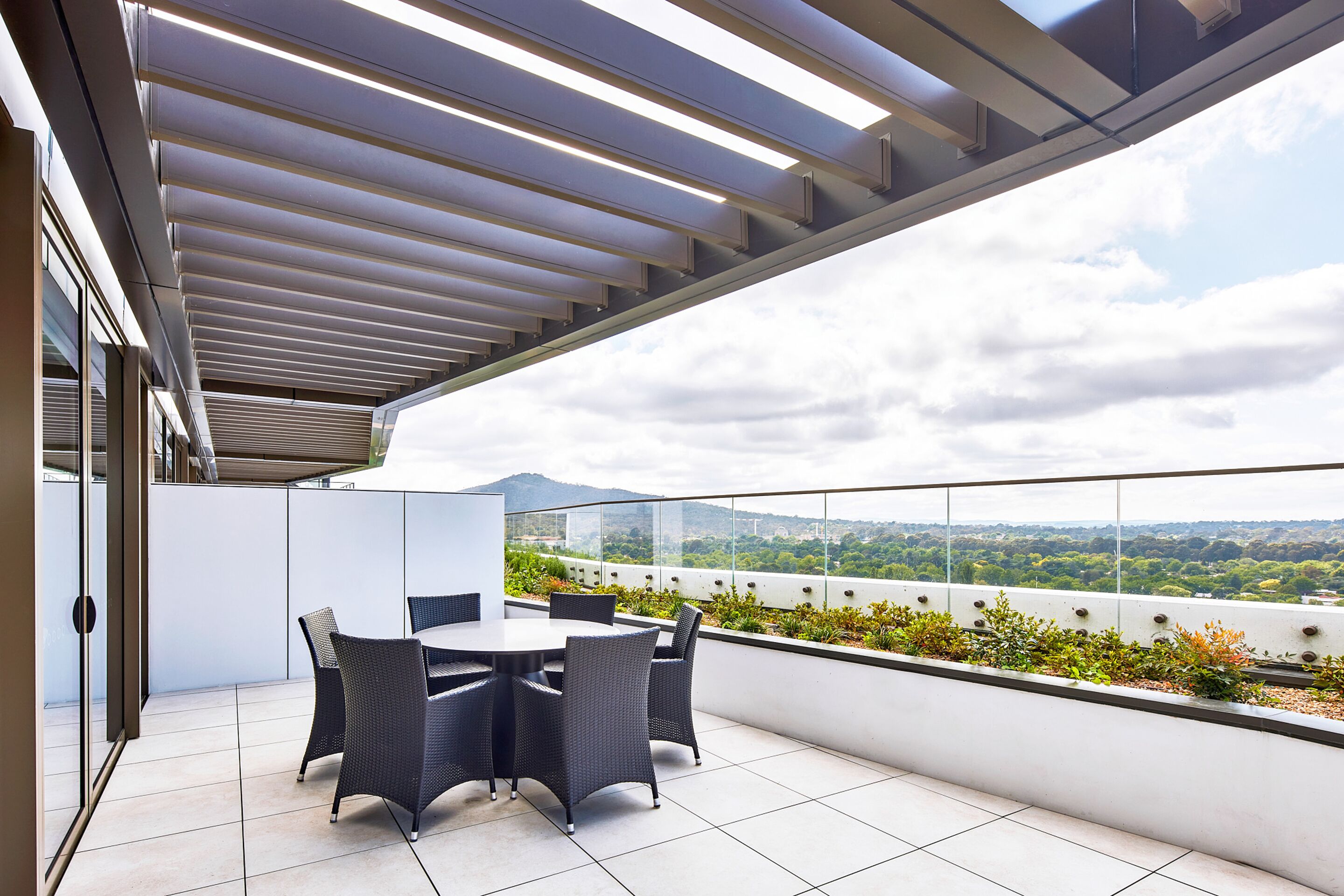 A rooftop terrace with a table and chairs, a glass railing, and a view of the cityscape.