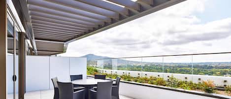 Una terraza en la azotea con una mesa y sillas, barandilla de vidrio y vistas panorámicas de la ciudad.
