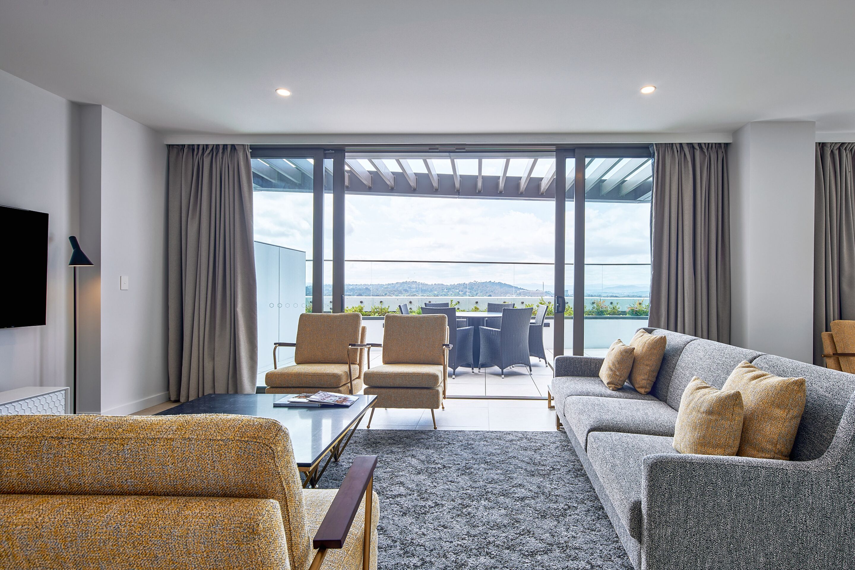En moderne stue med sofa, lænestole og sofabord. Et fjernsyn er monteret på væggen, og et stort vindue er dækket af gardiner.