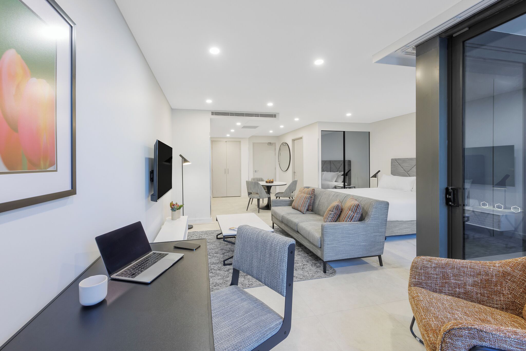 Ein modernes Hotelzimmer mit Schreibtisch, Laptop und Kaffeetasse. Ein graues Sofa und ein gemustertes Sessel sind vorhanden. Das Zimmer verfügt über ein großes Fenster und eine Glastür zum Balkon.