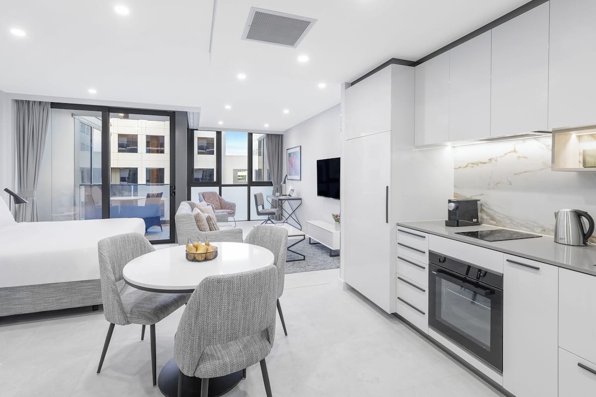 A modern hotel room with a dining area, kitchenette, and a large window offering a city view.