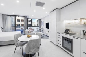 A modern hotel room with a dining area, kitchenette, and a large window offering a city view. - Meriton Suites Canberra (Canberra)