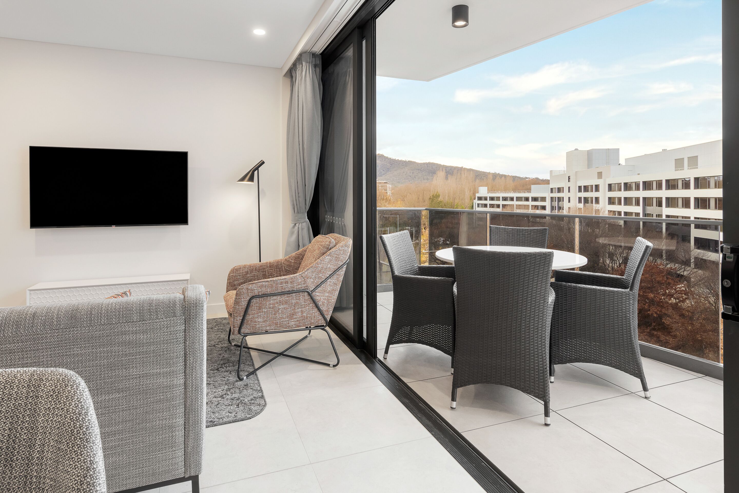 Una sala de estar moderna con un televisor de pantalla plana, sillones tapizados en gris, una mesa redonda y un ventanal con vistas a edificios y colinas.