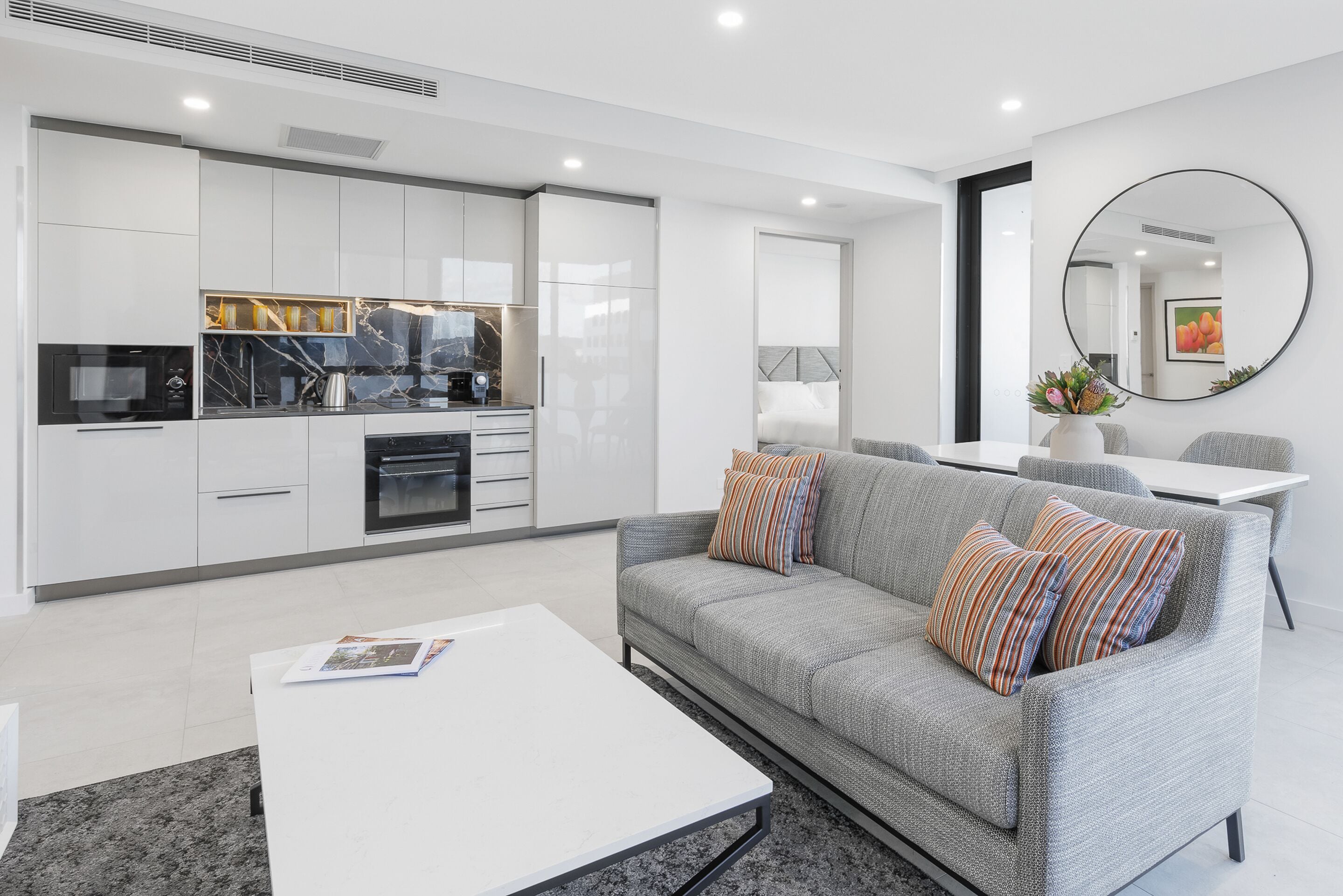 A modern living room with a grey sofa, white coffee table, and a kitchen area with white cabinets and a black countertop.