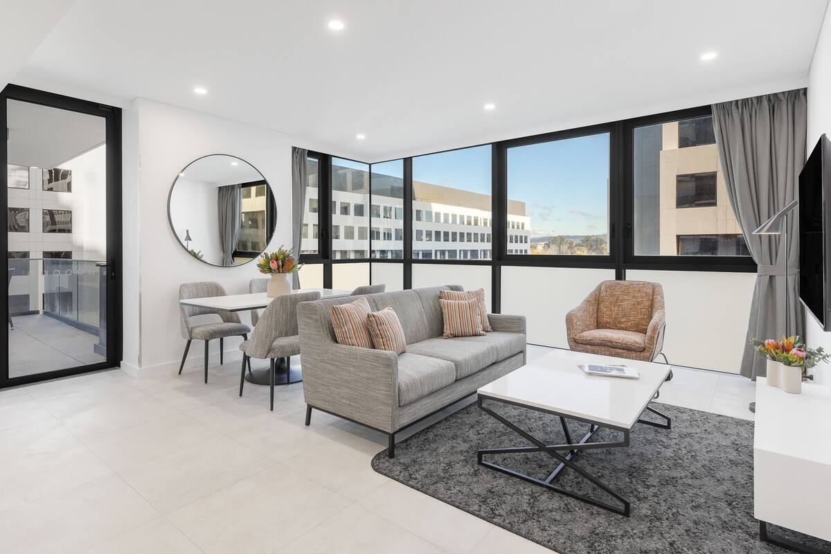 A modern living room with a grey sofa, a white coffee table, and a round mirror. There is a dining area with a table and chairs, and large windows with curtains.