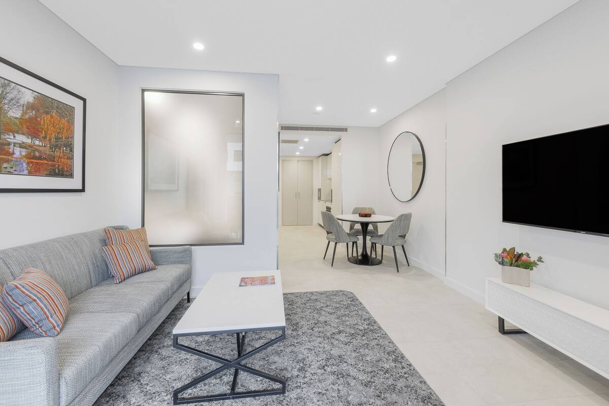 A modern living room with a grey sofa, a white coffee table, a dining area with a round table and chairs, a large flat-screen TV mounted on the wall, and a framed picture on the wall.