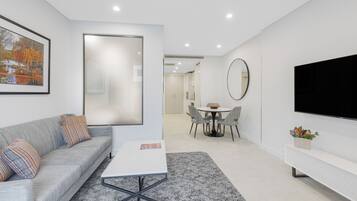 Uma sala de estar moderna com um sofá cinzento, uma mesa de centro branca, uma área de jantar com uma mesa redonda e cadeiras, uma grande TV de tela plana na parede e um quadro pendurado na parede.