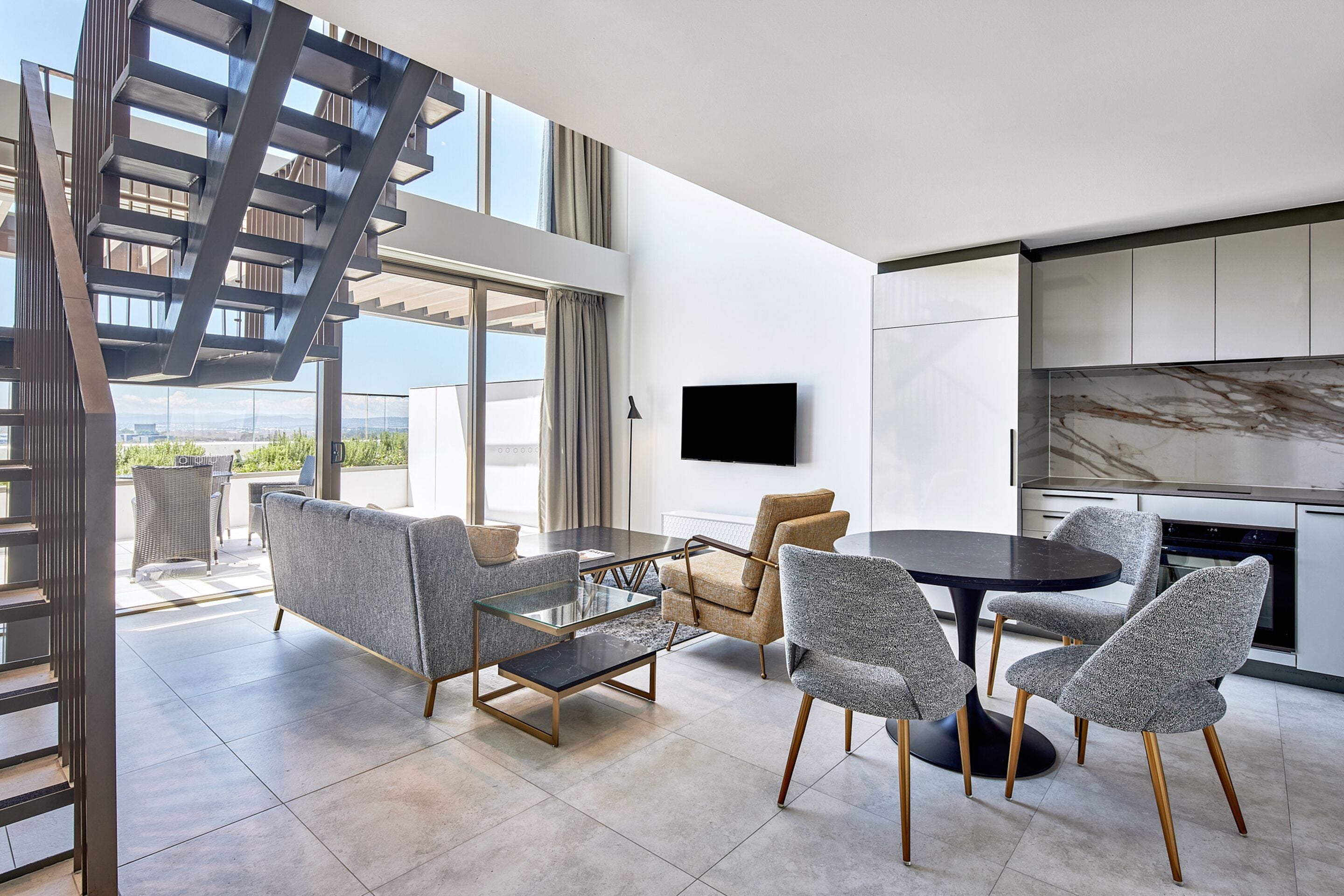 Un salon moderne avec une table basse en verre, des fauteuils à revêtement gris, une table ronde noire et un téléviseur à écran plat fixé au mur.