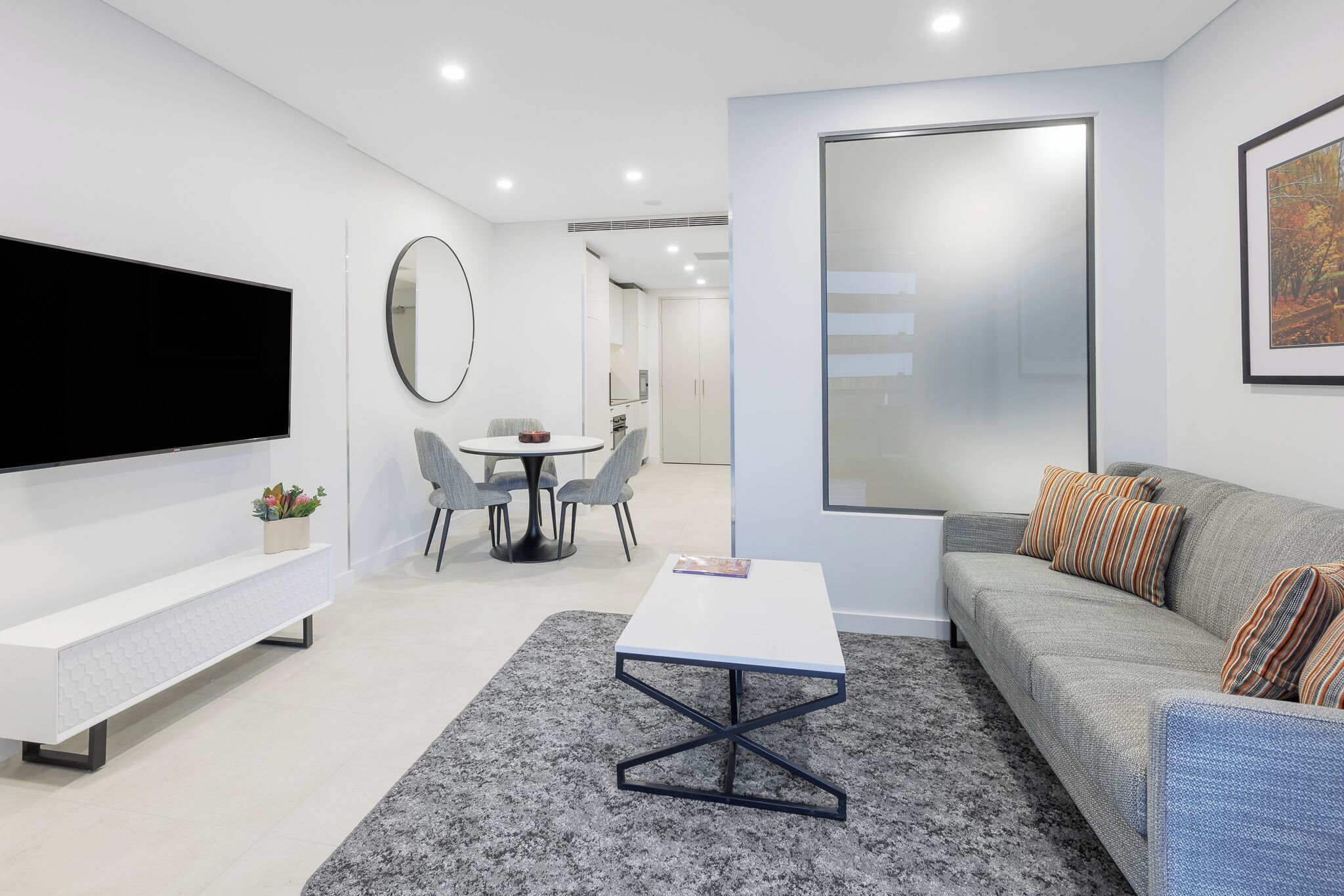 Una sala de estar moderna con un televisor de pantalla plana, una mesa de centro blanca, un sofá gris con cojines a rayas, y un comedor con una mesa redonda y sillas.
