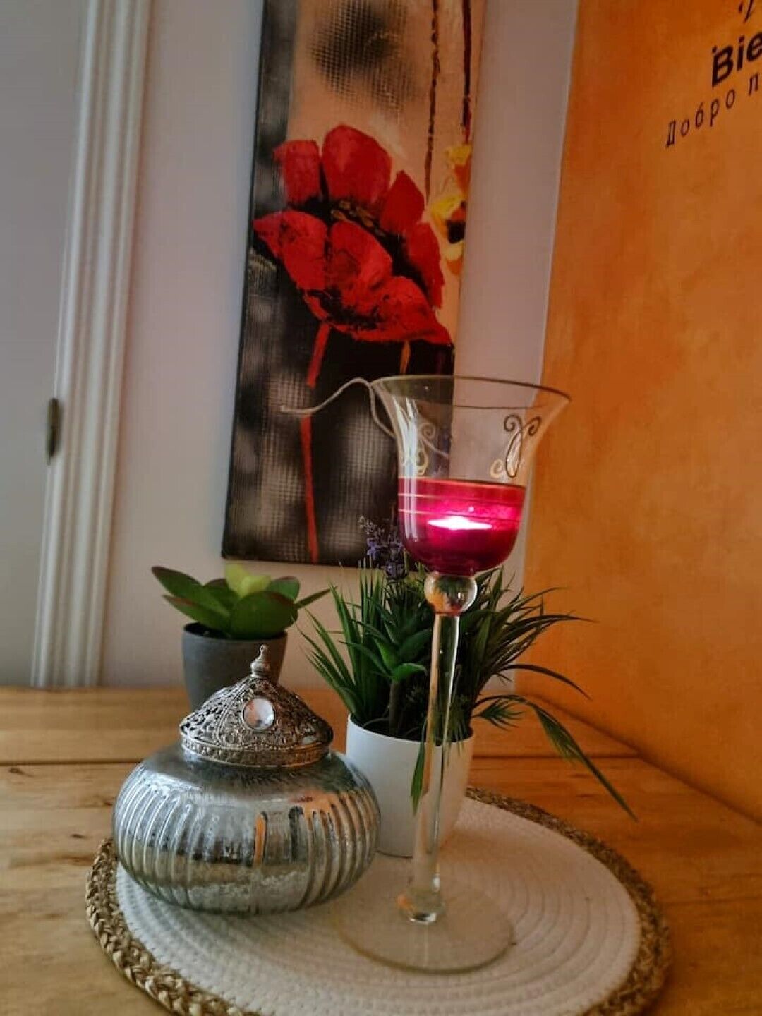 Un verre contenant un liquide rouge, un récipient en métal décoratif et deux plantes en pot placés sur un tapis tissé.
