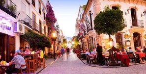 A narrow street lined with buildings, some with outdoor seating and tables, suggesting a dining area.