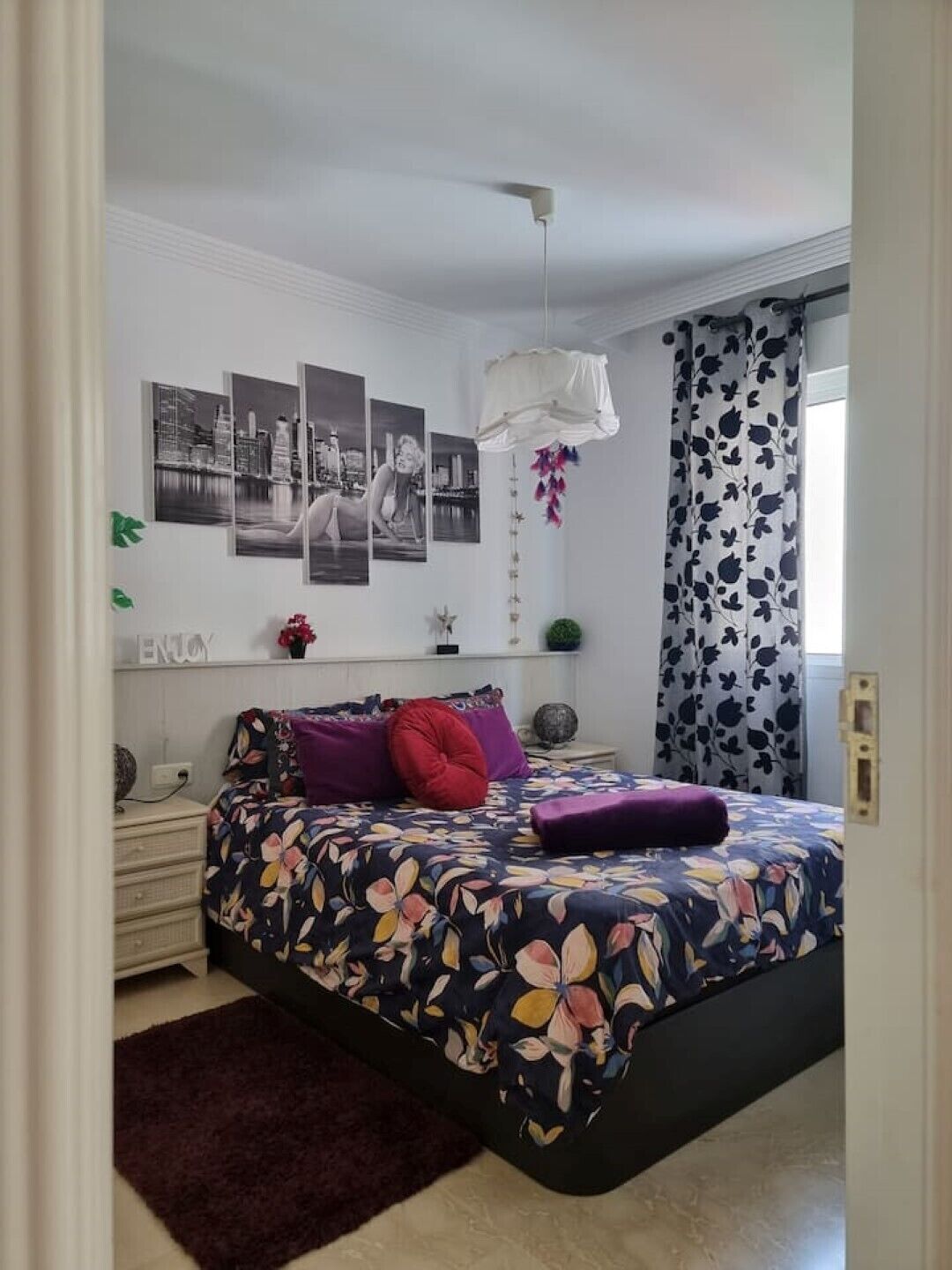Een slaapkamer met een bed, nachtkastje, kaptafel, raam met gordijnen en wanddecoratie.