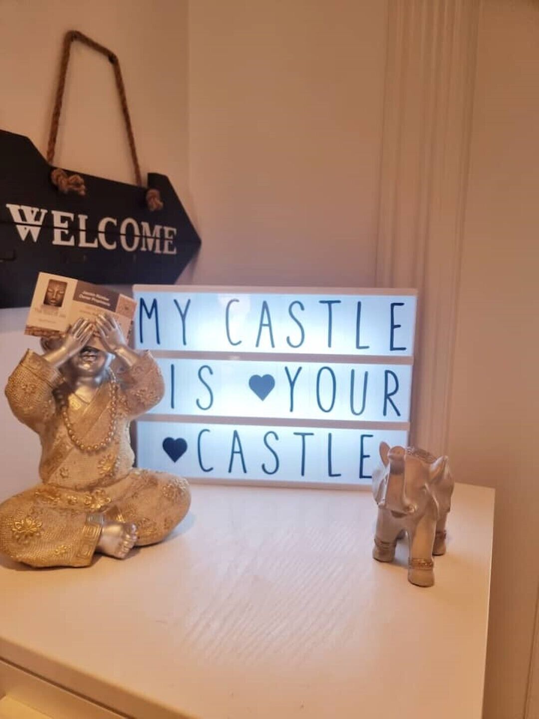 Eine dekorative Anordnung mit einem Schild, auf dem “MY CASTLE IS YOUR CASTLE” steht, und zwei Figuren auf einem Tisch.