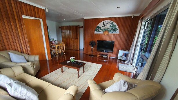 A spacious living room with a wooden floor, beige sofas, a coffee table, a TV, and a large window with curtains.