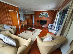 A spacious living room with a wooden floor, beige sofas, a coffee table, a TV, and a large window with curtains. - Spacious 4-bdrm fully self-contained WIFI, Parking (Suva)