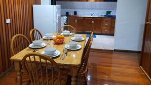 A dining area with a wooden table set for four, surrounded by wooden chairs. A basket of oranges is on the table. The kitchen in the background has wooden cabinets and a white refrigerator.