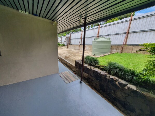 A covered outdoor area with a concrete floor, a stone retaining wall, and a grassy yard. - Spacious 4-bdrm fully self-contained WIFI, Parking (Suva)