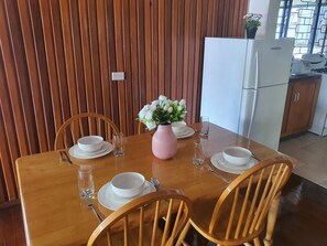 Una mesa de comedor dispuesta con platos, cuencos y cubiertos blancos, junto con un jarrón rosa con flores blancas.