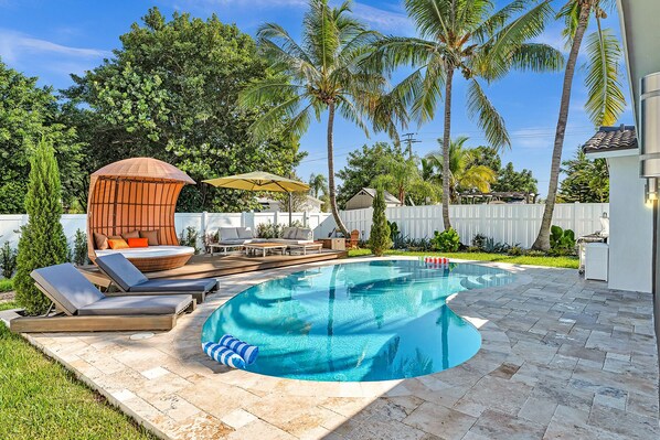 Un patio trasero con piscina, sillas de descanso, un área de estar techada y palmeras.