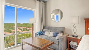 A bedroom with a large window, a grey sofa, a wooden table, a bedside table with a lamp, and a mirror on the wall. - Studio Sandestin Resort, 4 mins walk to beach, sleeps 3-4 (Miramar Beach)