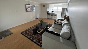 A modern living room with a sofa, coffee table, and a TV. There is a kitchen in the background with a refrigerator and cabinets. - Cheerful 3 bedroom  with free parking (Auckland)