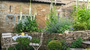Un muro de piedra rústica con una mesa y sillas, rodeado de abundante vegetación y plantas con flores.