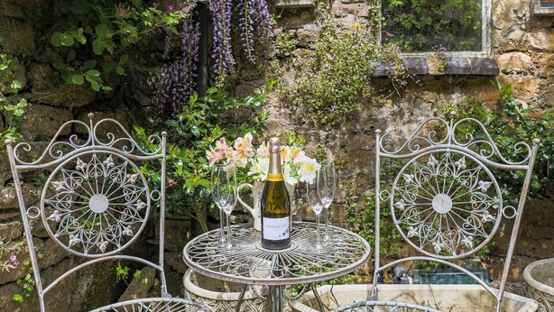 Una mesa de metal con una botella de vino y dos copas, rodeada de sillas de metal decoradas, frente a una pared de piedra con follaje y flores moradas.
