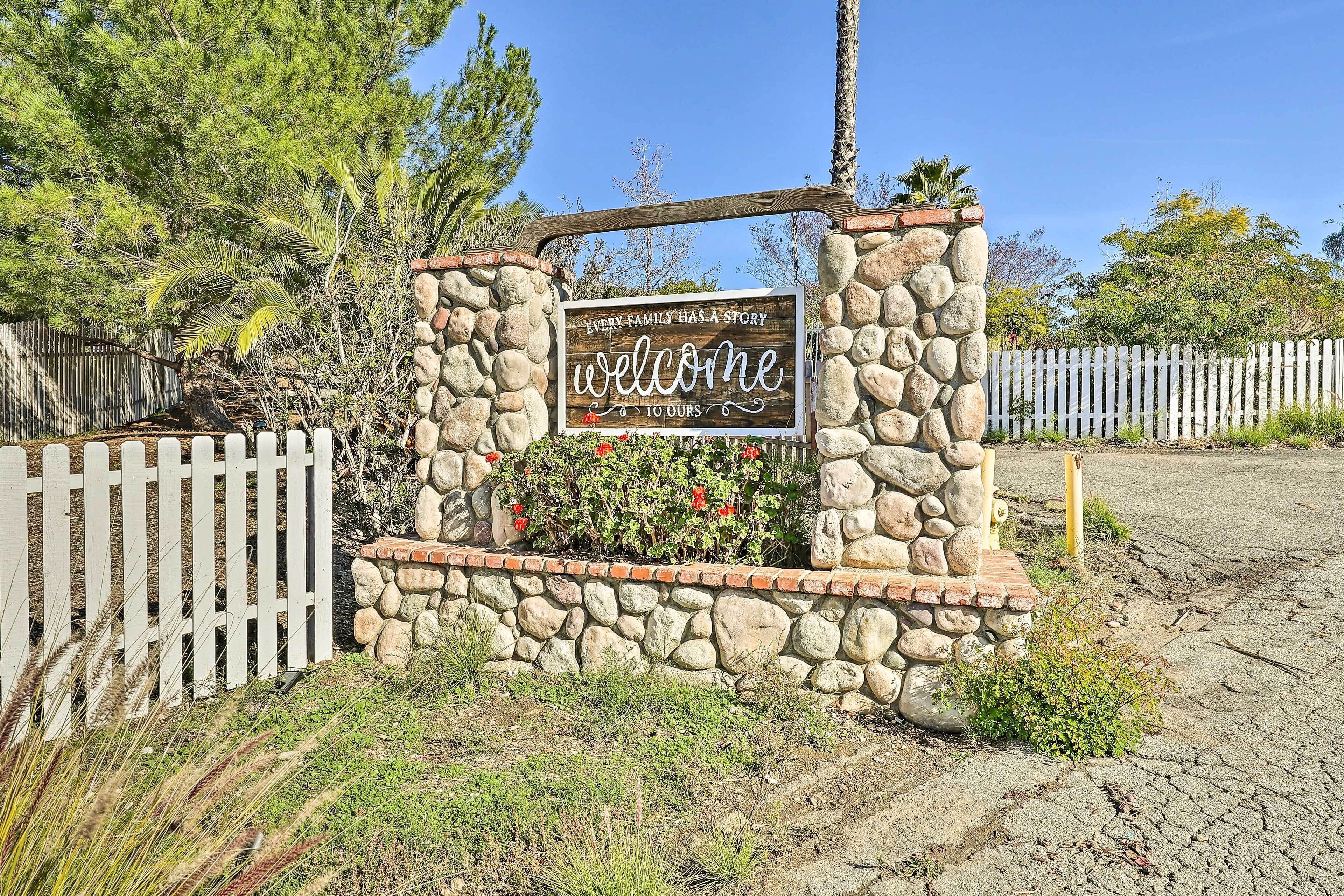 A welcome sign with a stone and brick border, surrounded by a white picket fence and greenery.
