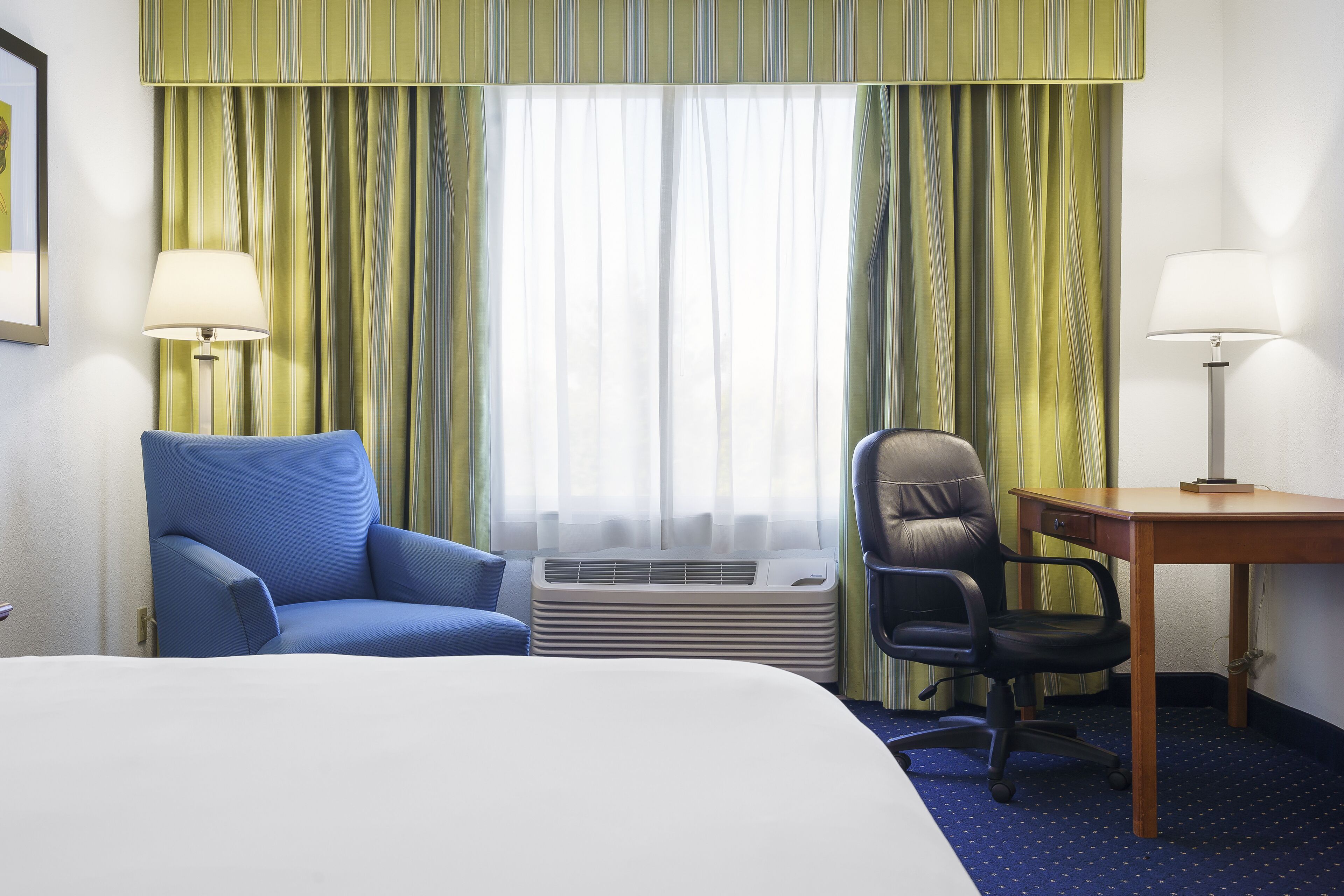 Una habitación de hotel con una cama, un sillón azul, un escritorio de madera, una silla de oficina negra, una lámpara de pie y un equipo de aire acondicionado.