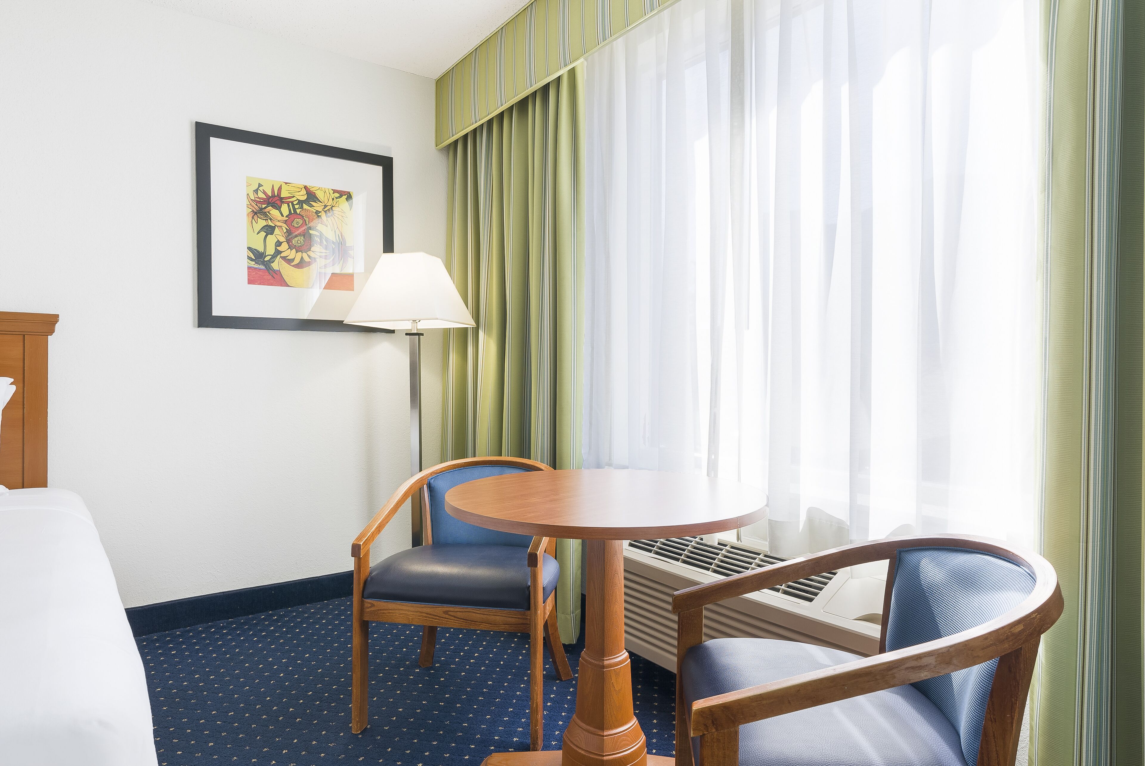 A hotel room with a bed, a desk with a lamp, two chairs, a round table, and a window with curtains.
